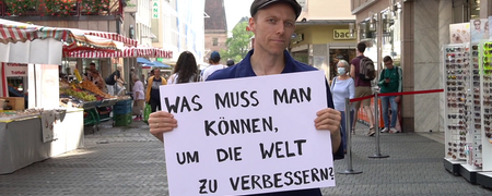 Ein Mann steht in der Fußgängerzone mit einem Schild: "Was muss man können, um die Welt zu verbessern?"  - Link auf: Selbsttest