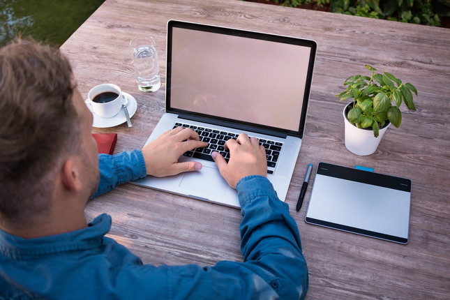 Ein Mann tippt im Homeoffice an seinem Laptop, neben ihm stehen ein Kaffee, ein Tablet und eine Tischpflanze