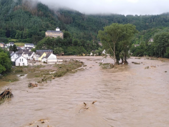 Die Aufnahme zeigt ein Tal, links stehen Häuser und mit Bergen im Hintergrund, das überschwemmt ist. 