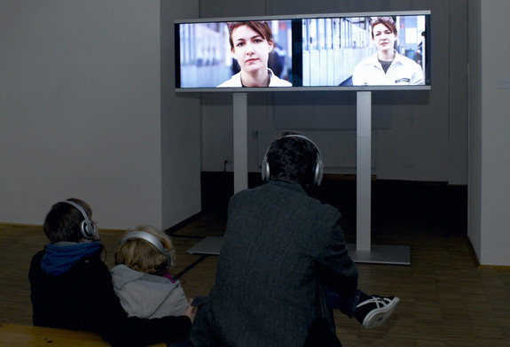 Das Bild zeigt eine Videoinstallation, vor der drei Menschen sitzen