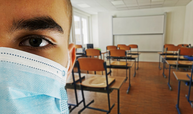 Junge mit Mund-Nasen-Schutz vor leerem Klassenzimmer mit hochgestellten Stühlen