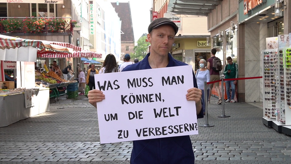 Ein Mann steht in der Fußgängerzone mit einem Schild: "Was muss man können, um die Welt zu verbessern?"