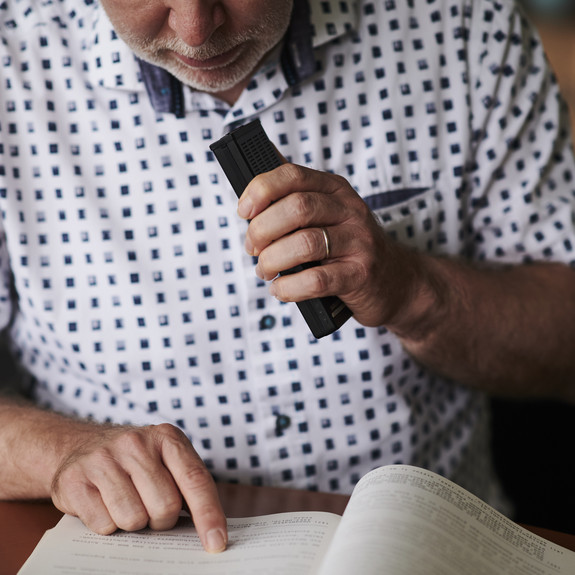 Ein Mann hat ein Diktiergerät in der Hand, das er an seinen Mund hält. Der Zeigefinger der anderen Hand liegt auf einer Zeile eines aufgeklappten Buches.