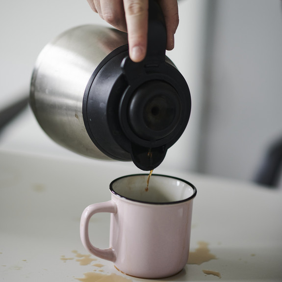 Jemand schenkt Kaffee aus einer Kanne in eine Tasse. Um die Tasse sind Kaffeeflecken auf dem Tisch.