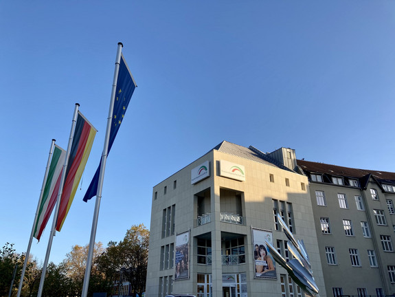 Das Bild zeigt das Gebäude der Auslandsgesellschaft in Dortmund. Vor ihm stehen drei Fahnenmasten mit Flaggen von Deutschland, Nordrhein-Westfalen und der Europäischen Union.