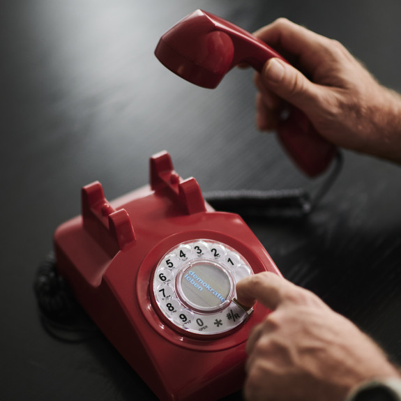 Ein rotes Telefon mit Wählscheibe steht auf einem dunklen Tisch. Jemand hat den Hörer in der Hand. Die andere Hand wählt eine Nummer.