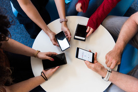 Das Bild zeigt einen Tisch von oben sowie vier Armpaare mit vier Smartphones.