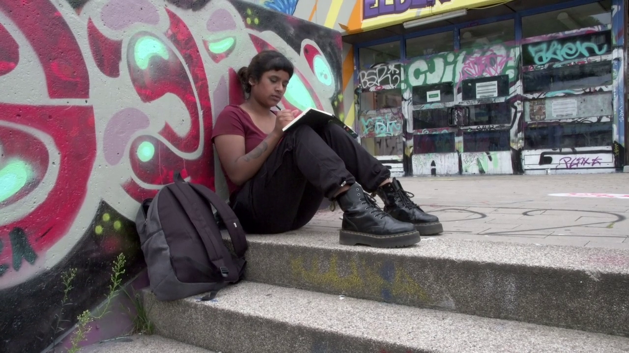 Eine junge POC-Frau sitzt an der Mauer und zeichnet in ihrem Heft